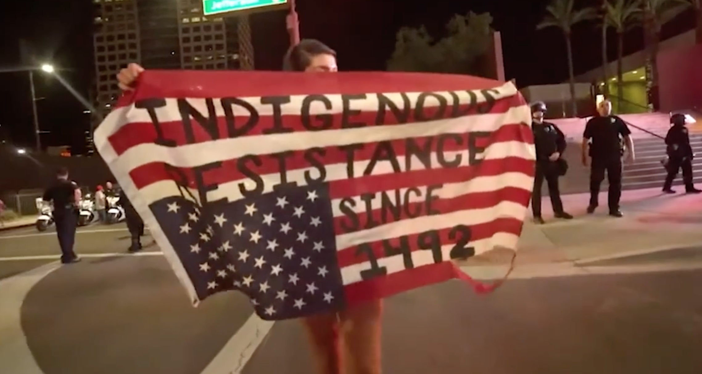 Protestor carrying an upside-down US flag marked with the words “Indigenous resistance since 1492.”