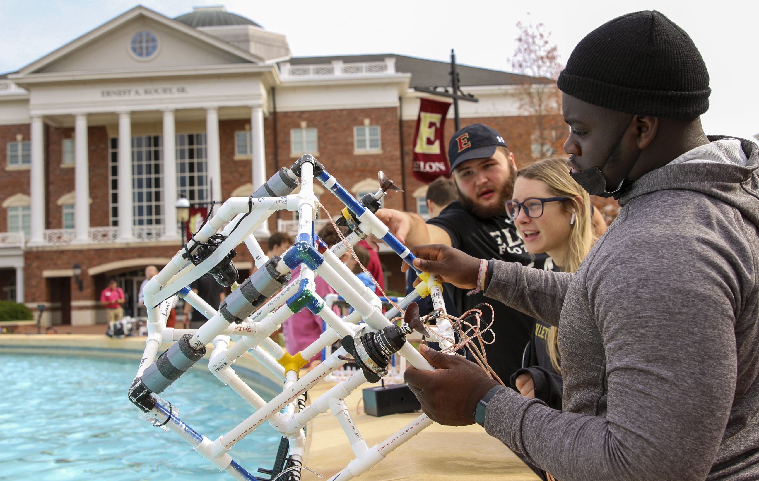 Three students examine their underwater remotely operated vehicle prototype made from PVC pipe, rake tines and pool noodles in an outdoor fountain on Elon's campus.