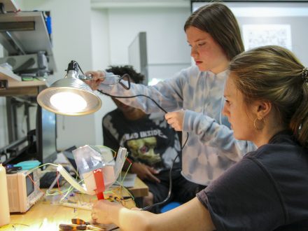 Two female students building a small, solar-powered electronic device and holding a large lamp over its sensor.