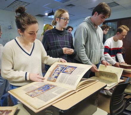Delaney Guidi '26 and other classmates surround and flip through large books of colorful and intricately detailed medieval art facsimiles in a classroom.
