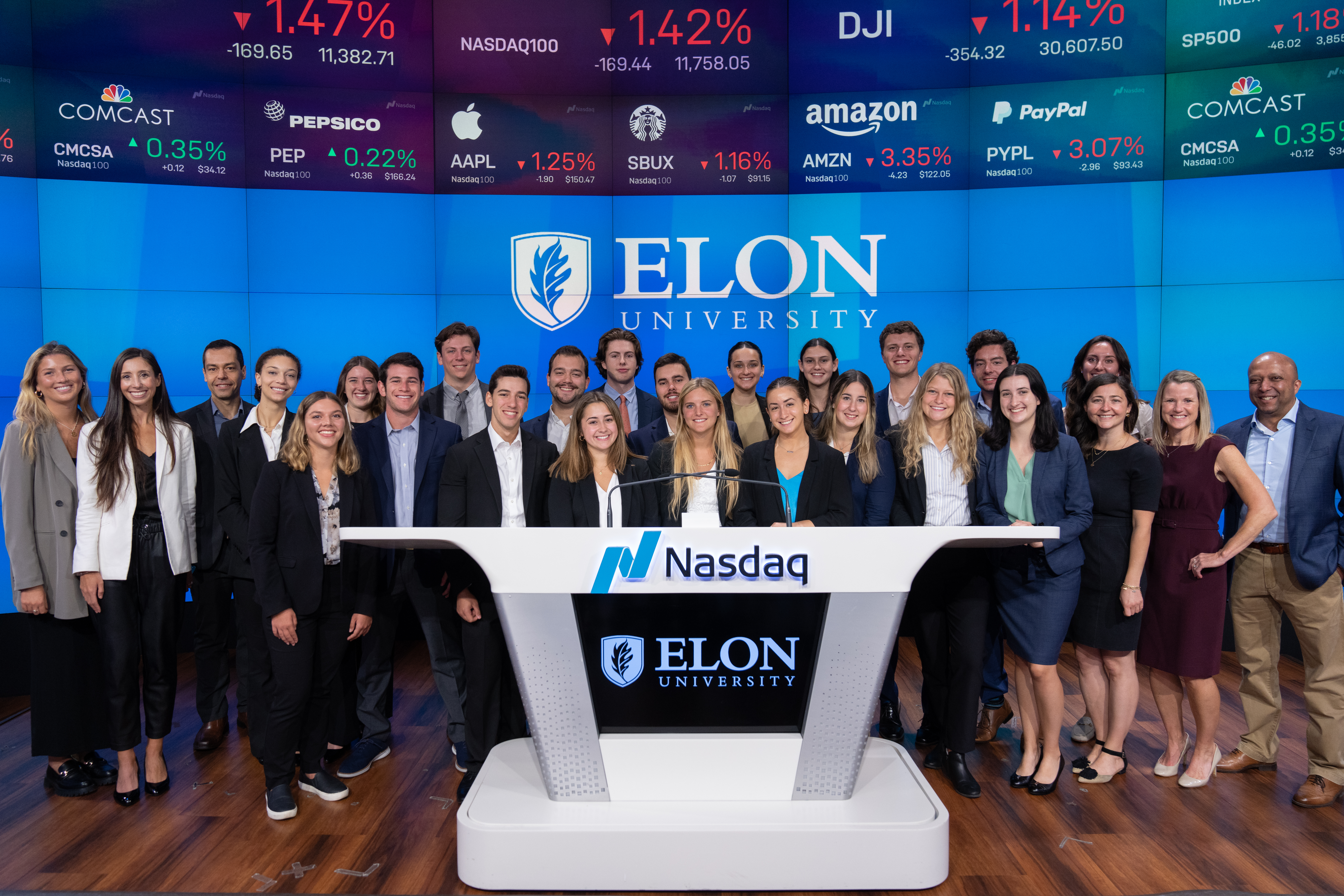 Group of students, faculty and staff standing beneath stock signs and behind lectern that says Nasdaq and Elon University