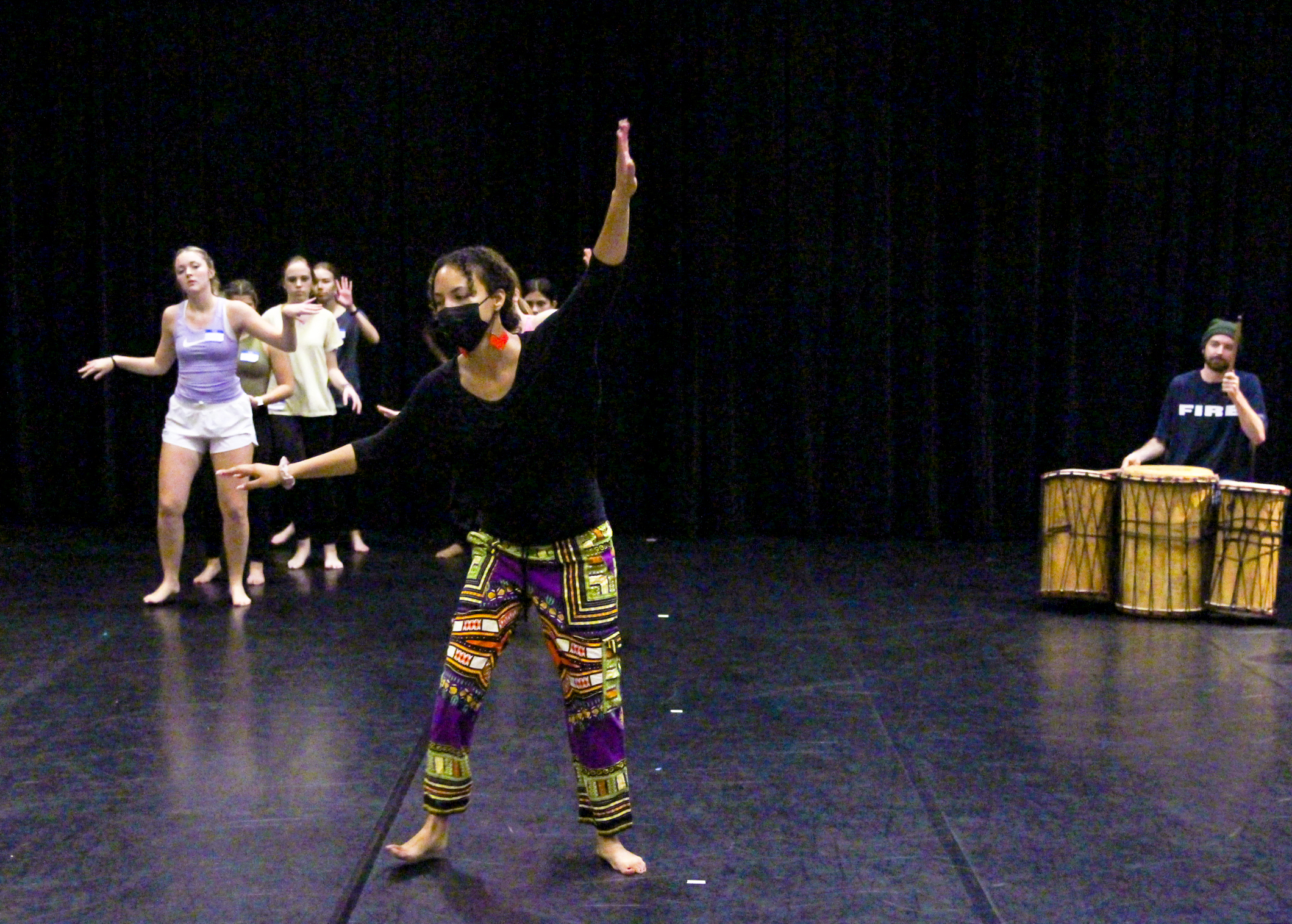 A professor demonstrates movements associated with West African dance for students standing in two lines behind her while a man at the side of the Roberts Theatre stage keeps rhythm on a set of drums.