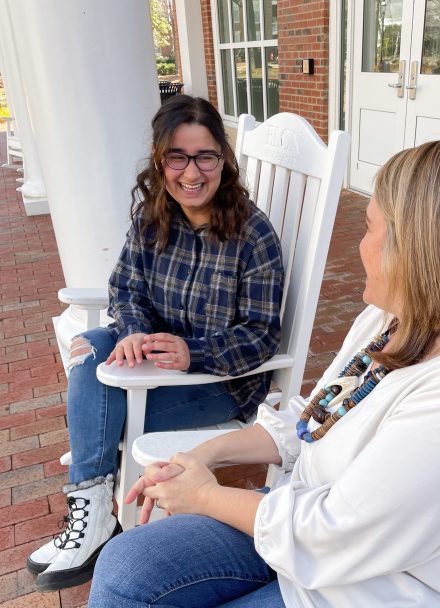 female student wearing glasses sitting in a white rocking chair talking with a female professor outside