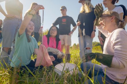 A group of students and a professor in a sunny field gathered around a a student as she weighs a juvenile snake inside of a specimen bag.