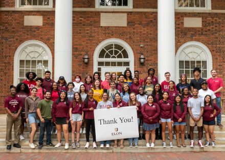 A group of Odyssey Scholars stand in front of Whitley Auditorium at Elon University holding a "thank you" sign.