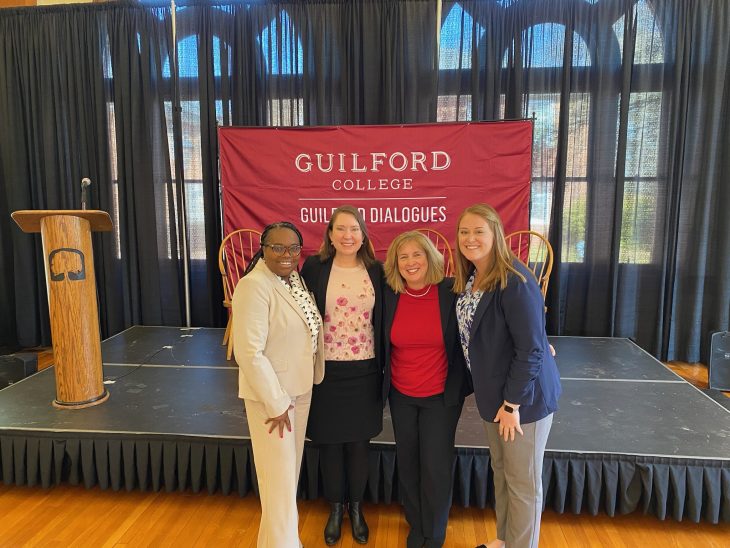One Black woman standing beside three white women in front of a Guilford College maroon banner