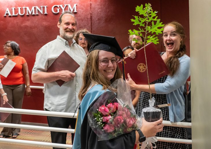 A graduate poses with flowers, her sapling and her family outside Alumni Gym