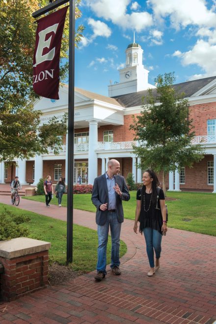 A professor and student chat as they walk along the brick walkway in front of Lindner Hall on Elon's campus.