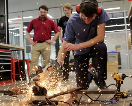 A student using a saw to cut metal, throwing a shower of sparks onto the floor of the prototype lab
