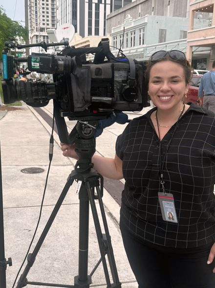A female student stands with a large camera while shooting on sidewalk in South Florida.