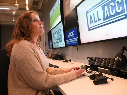 Female student sits in ACC game day operations room surrounded by monitors.