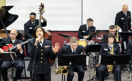 A group of musicians sing and play wearing US Navy dress uniforms.