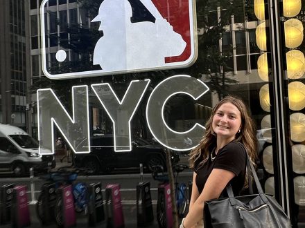 Emilia Cappellett smiles in front of a MLB sign located against a wall of glass.