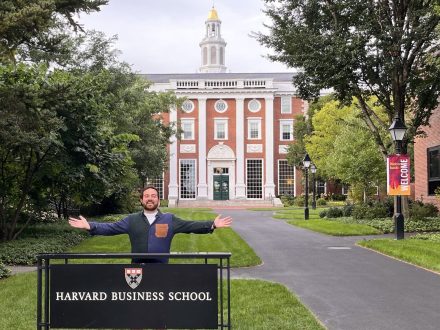 Matt Sears with arms outstretched at the Harvard Business School sign with the building pictured behind him