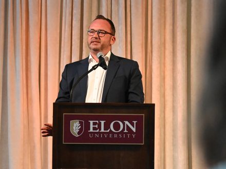 A white male stands at a podium with Elon University on the sign in front of him.