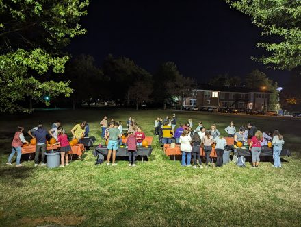 Large lawn at night with students gathering around multiple tables carving pumpkins