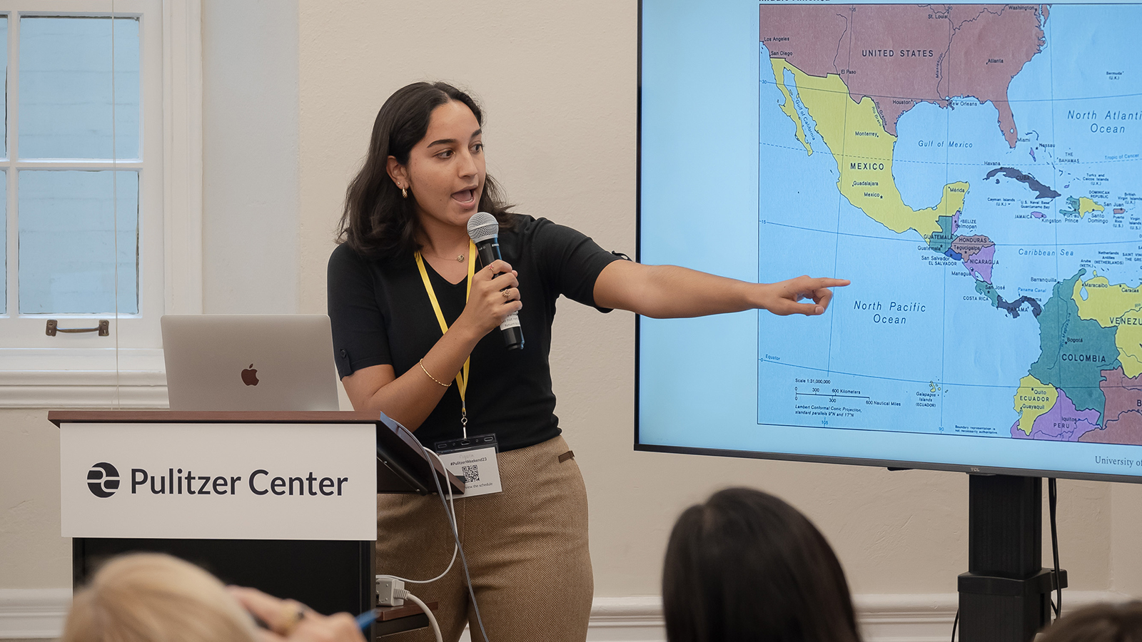 A young female reporter stands at a branded Pulitzer Center podium, pointing to a map of Central America.