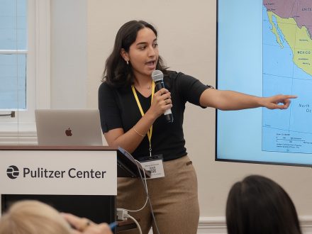 A young female reporter stands at a branded Pulitzer Center podium, pointing to a map of Central America.