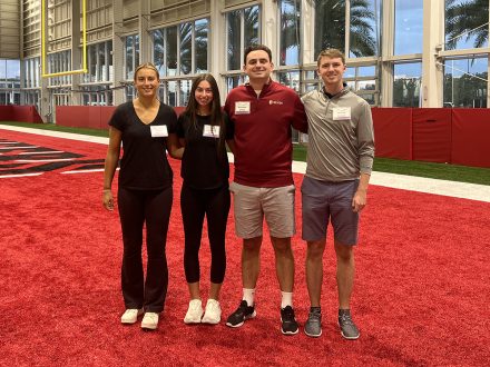 Four students stand on a red end zone in an indoor football complex.