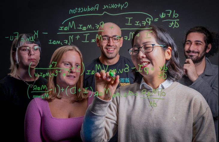 View through a glass board of a professor writing an equation with four students looking on