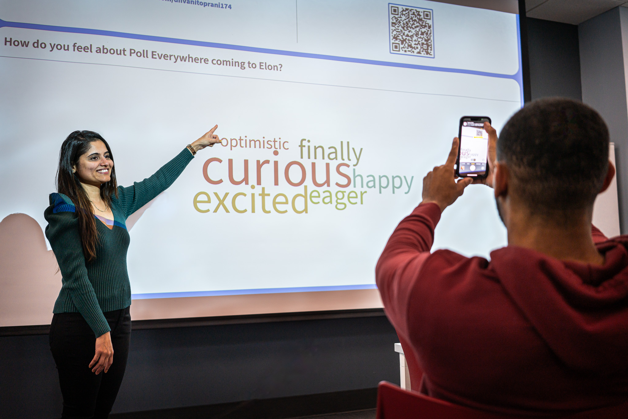 Class instructor with black hair wearing a dark green v-neck sweater standing on the left points to QR Code on the upper right of projector screen while a student wearing an Elon red hoodie scans the QR code with their smartphone. In the middle of the projector screen is a word cloud reading 