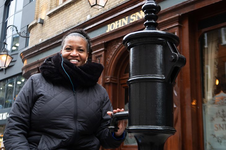 Callie Kelly stands in front of the Broad Street Pump. The pump is as tall as she is and features a matte black iron surface. Kelly has her hand on the handle and is wearing a large black coat. Behind her you can see the front of the John Snow Pub.