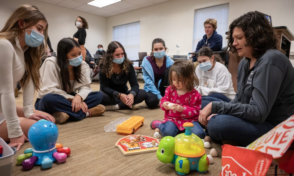 A woman and toddler play with toys as students watch