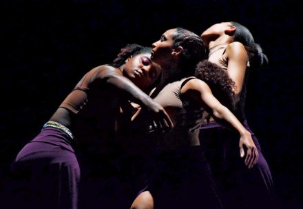 Three Black women dancing against a dark background.