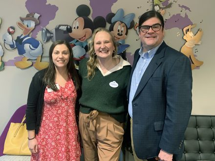 Two Elon University staffers stand with an Elon alumna in front of Disney sign.