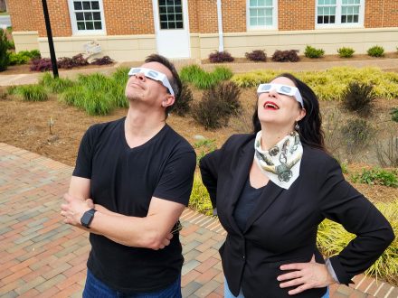 A man and woman wearing eclipse glasses and looking toward the sky