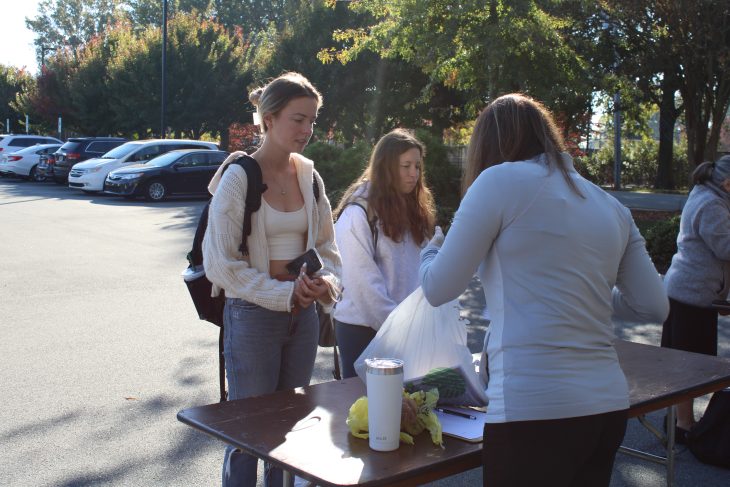 A student in a white shirt wearing a pony tail stands outside by a table as someone in. front of her is looking through a plastic bag with supplies