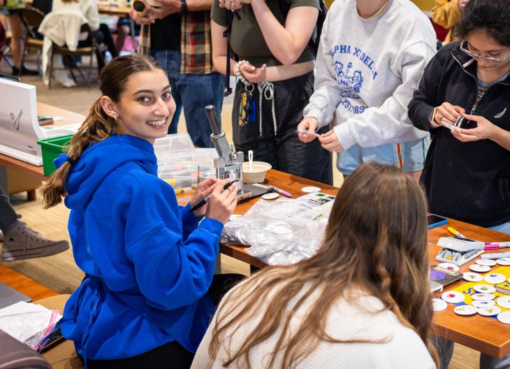 Maker Hub Consultant (female student with brown hair, green eyes, wearing a blue hoodie and apron) smiles at the camera over her left shoulder while sitting at sticker and button table. There is a line of standing students waiting to make their crafts in front of her.