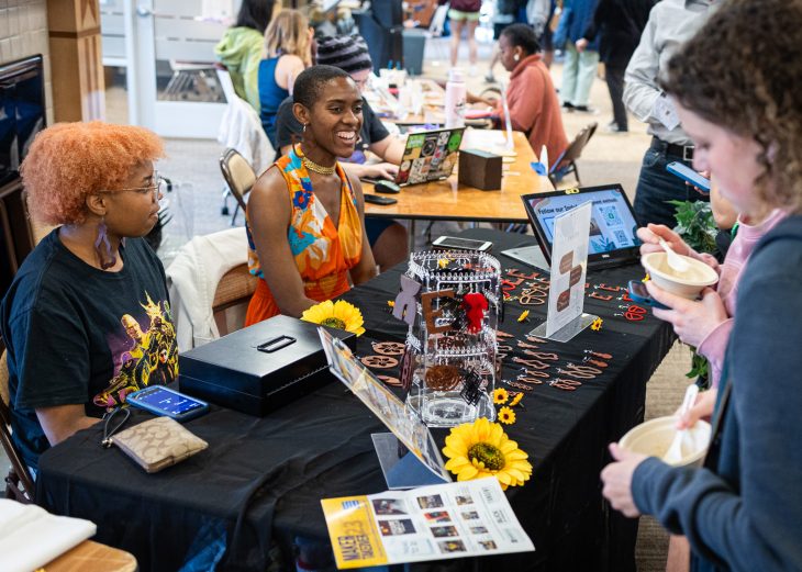 Two smiling female students sit behind their table that has sunflower heads and hand-made earrings while they talk with guests at their booth