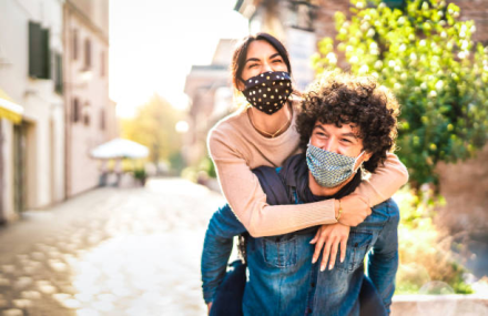 A woman on the back of a man, with both in masks. They are outdoors.