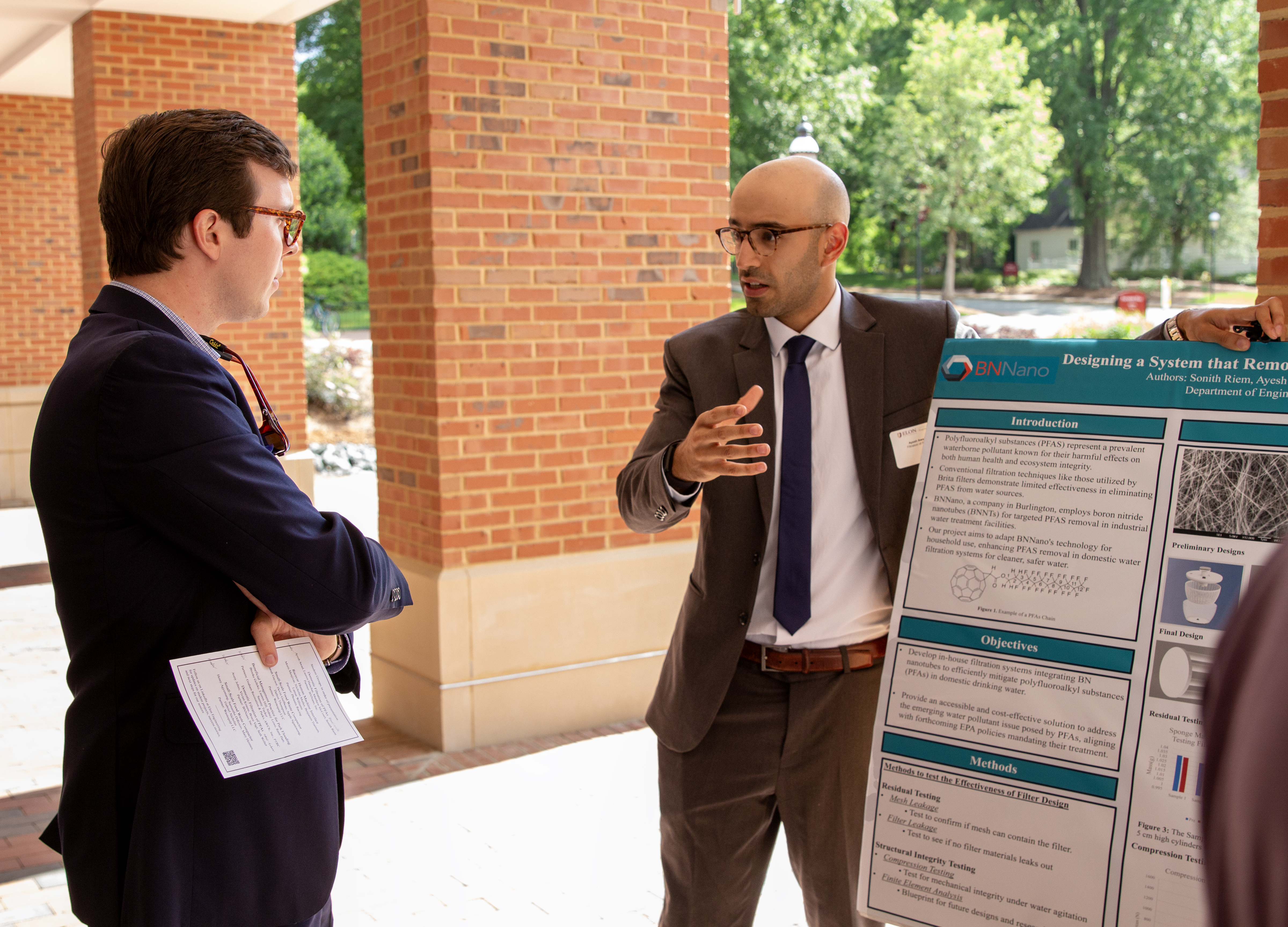 A student dressed in a suit speaks with a man n front of a research poster 