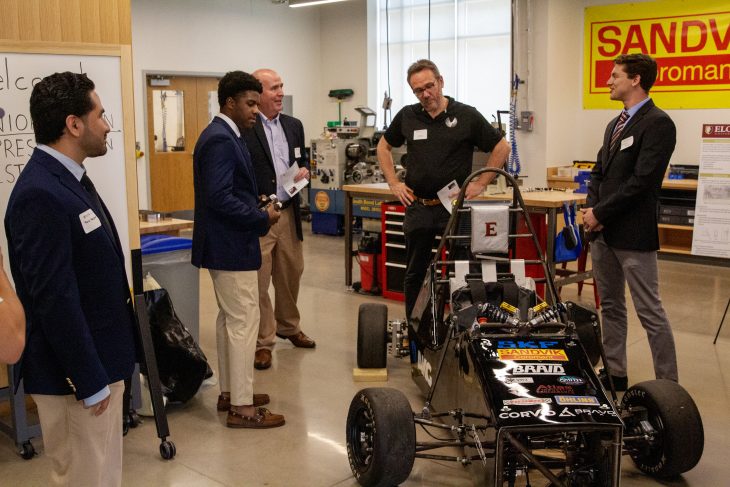 Three students and two men stand around a race car in a machine shop