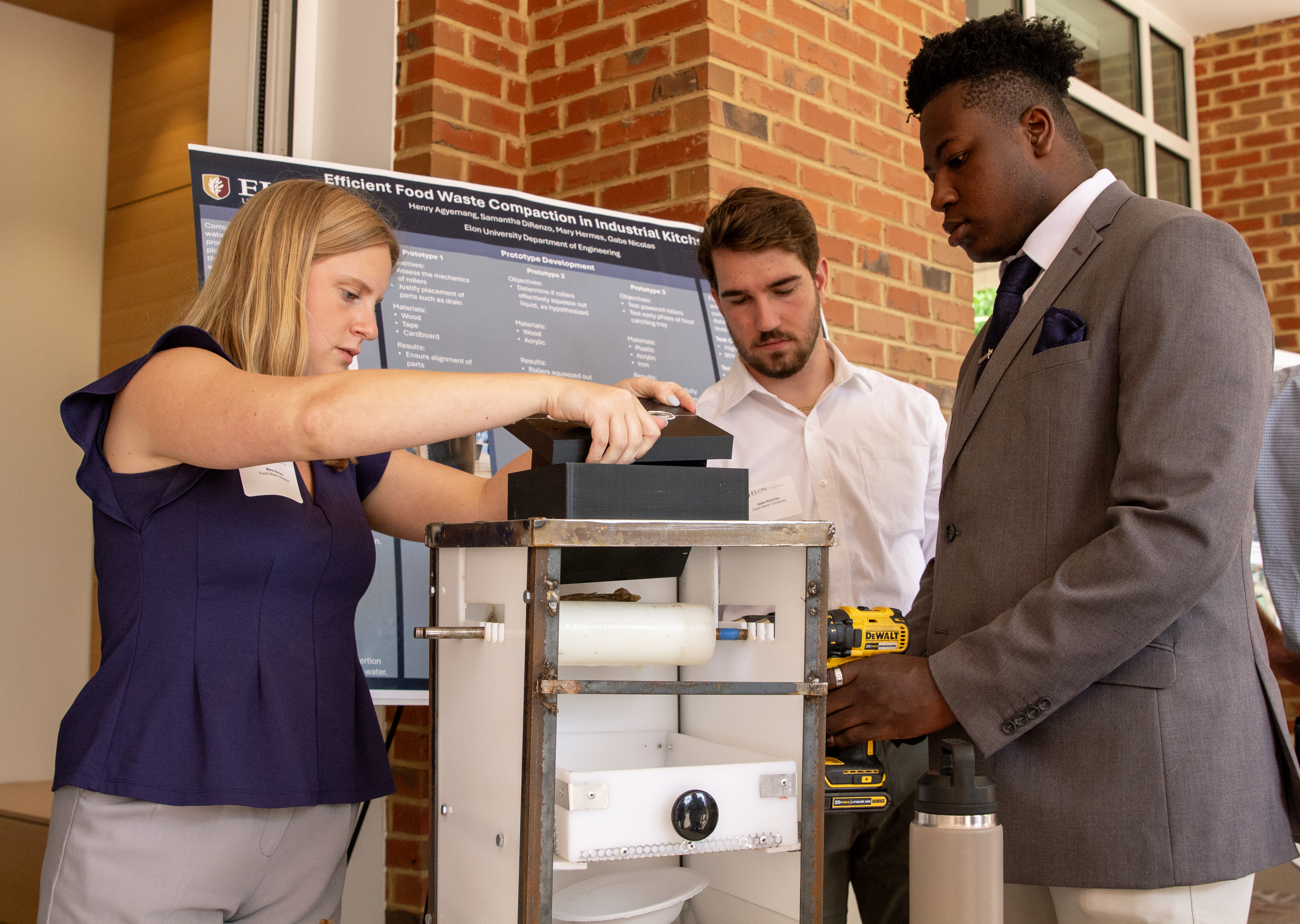 Three students around a table-top machine that processes food for composting in front of Founders Hall