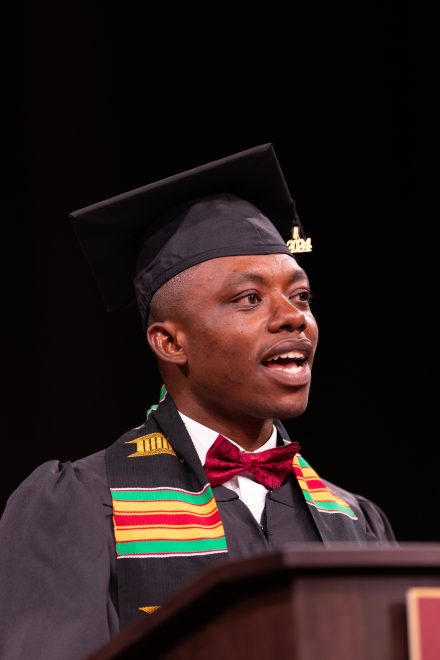 A male graduate in a cap and gown speaks at a podium