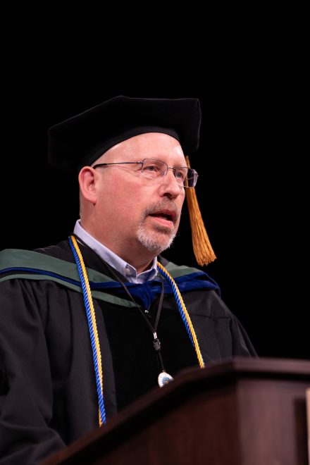A male commencement speaker in academic regalia stands at a podium