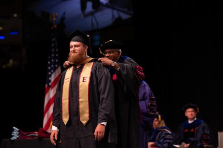 A male graduate in a cap and gown is hooded by a male professor in academic regalia