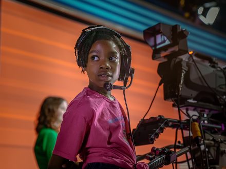 A young Black girl looks back while working a studio camera.
