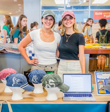 Two female students stand behind their table of custom designed hats,. The table has a banner with their business name R'KIND. On top of the table are four hats ine with a dragon, one with a tiger, one with a horse, and one can't be seen. Both students are wearing hats. The student on the left wears a white tee with a green hat with a tiger on the side. The student on the right is wearing a black crop top and red hat with a serpentine dragon on the side.