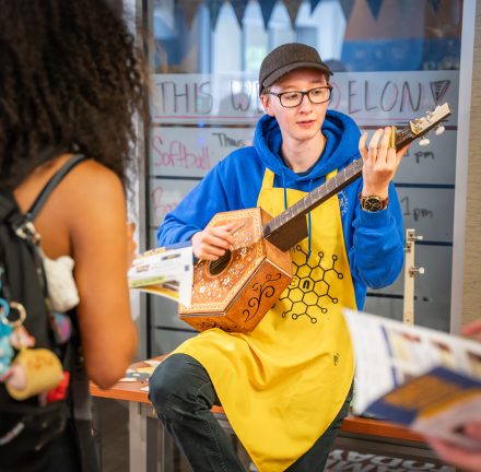 Student wearing blue hoodie, yellow Maker Hub apron, black rimmed glasses, and a grey hat strums a guitar shaped wooden instrument with a hexagonal body, while leaning backwards against a table for a female student with textured, shoulder-length hair