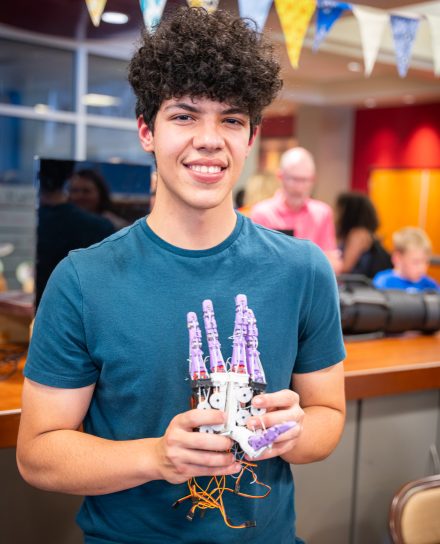 Male student with teal tee and dark curly hair shows a robotic hand, with purple 3D printed fingers, to the camera