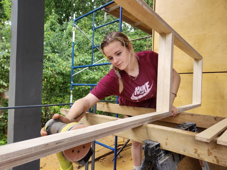 A woman in a maroon shirt uses an electric sander on a wooden frame