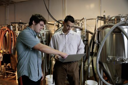 Two people look at a laptop screen with beer brewing equipment behind them