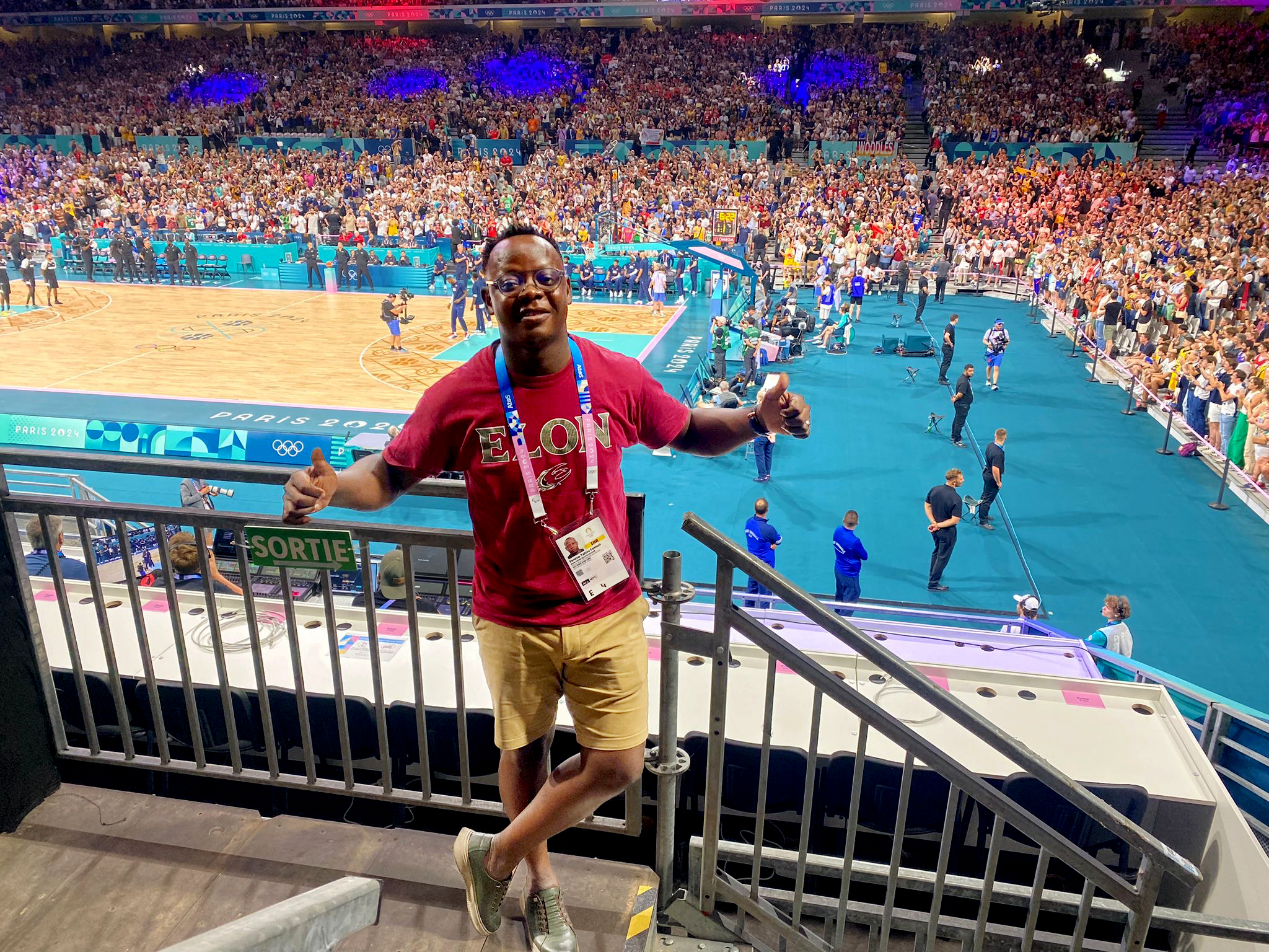 A man standing in a sports arena wearing an Elon shirt giving the thumbs up