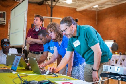 A group of people are bent over looking at silver macbook laptops with their hands navigating the trackpad. The most in-focus person is a man on the right facing the left, wearing a teal polo shirt and khaki pants and glasses. He has brown hair pulled back into a bun and black framed glasses.