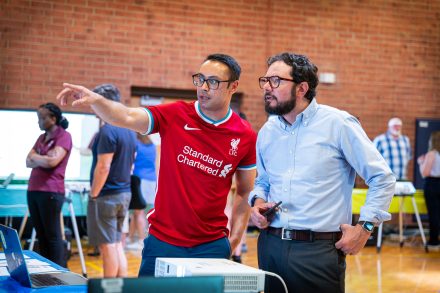 A Nepali man with black hair in a red Liverpool soccer jersey points off camera to a projector screen while a white man with wavy dark brown hair, a full goatee beard, glasses, and a blue button-down shirt looks on