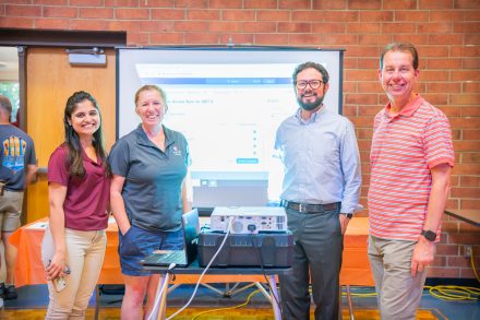 Four people stand in front of a projector screen with a brick wall behind it. The four people from left to right: Dhvani Toprani (an Indian woman with straight black hair wearing a maroon polo with khaki pants), Claudia Sparks (a white woman with red wavy hair, pulled back into a ponytail, wearing a gray polo and blue shorts), Michael Gartner (a white man with curly dark brown hair and glasses, wearing a blue button down and gray slacks), and Christopher Waters (a white man with medium brown hair, wearing a red, white, and blue, striped shirt and khaki pants).